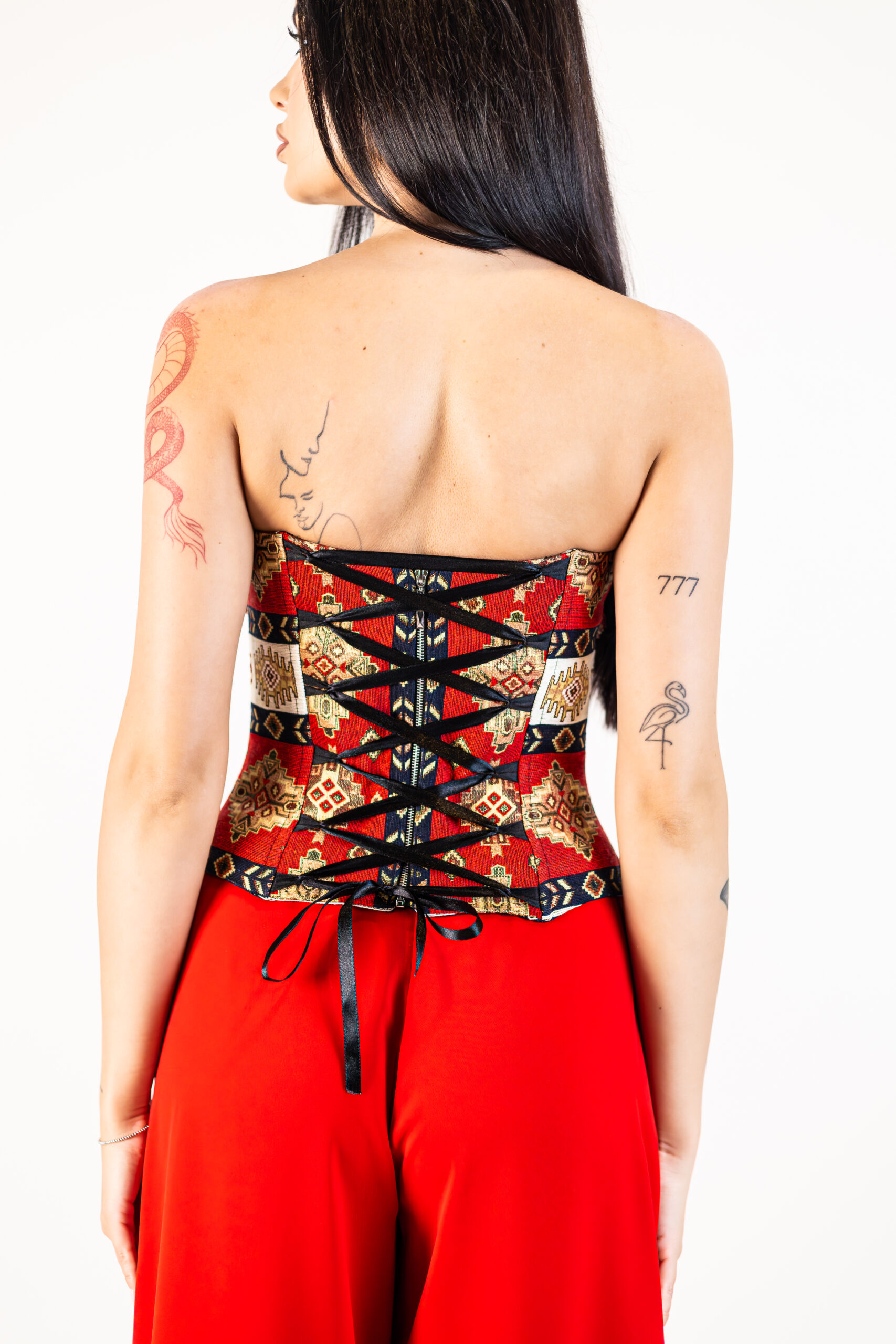 Corset Tessina Roșu din brocart fără bretele, cu motive geometrice și șnur reglabil din satin la spate, purtat cu pantaloni roșii eleganți.
